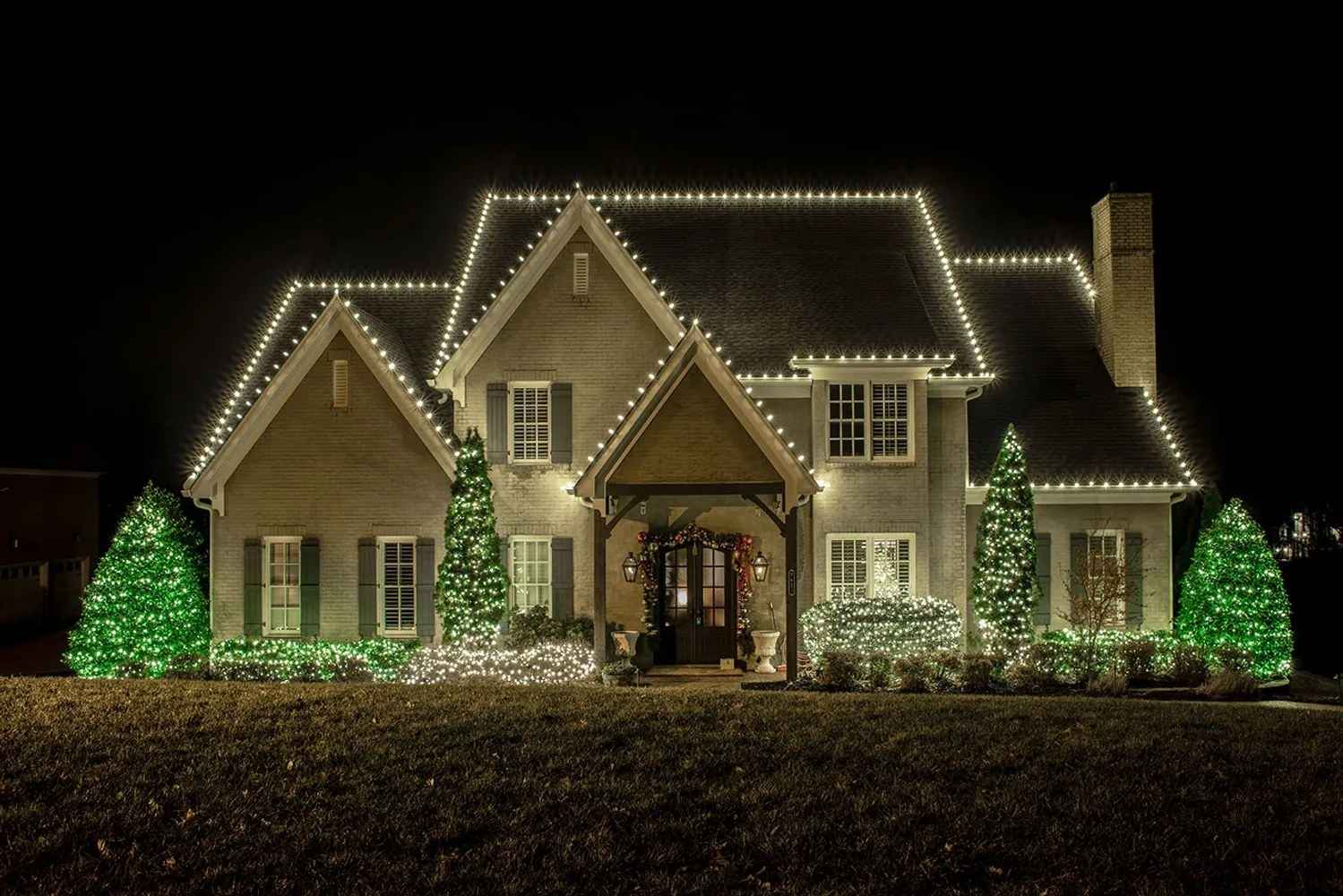 Home at night with white C9 lights on every roofline and green-lit evergreens out front