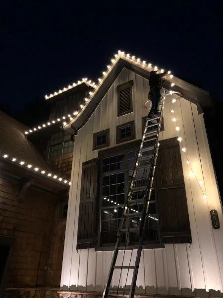 Installer on a tall ladder hanging white C9 lights along a gable at night