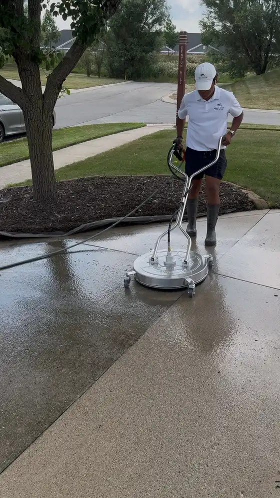 Technician running a round surface cleaner over a wet concrete driveway beside the lawn