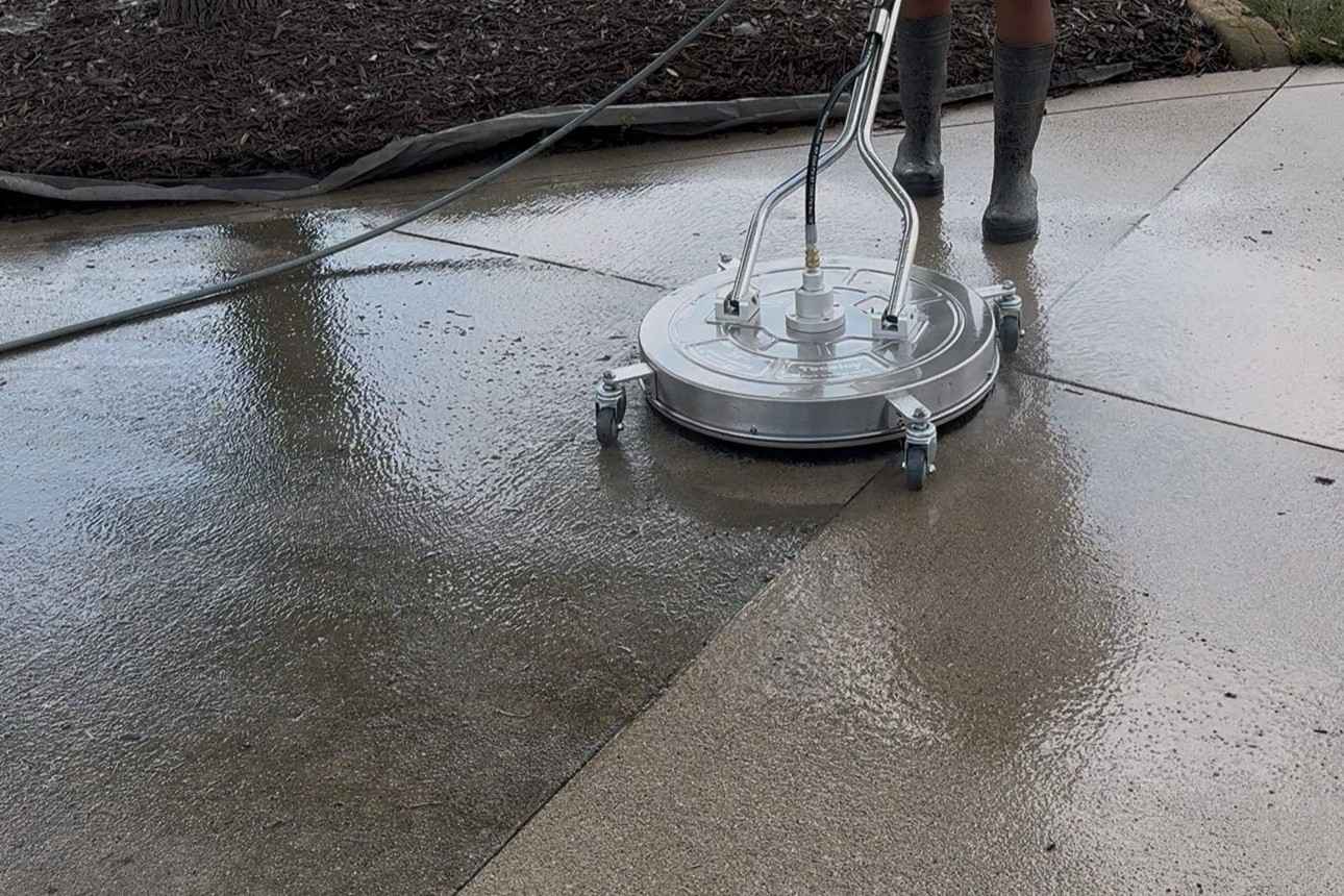 Round commercial surface cleaner pressure washing a concrete driveway, leaving a clean wet path