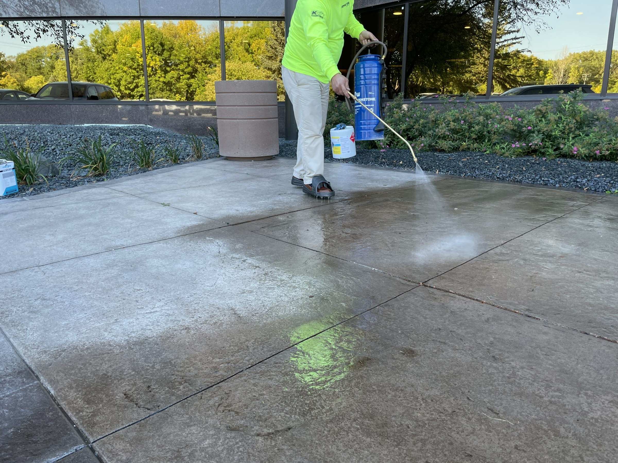 Worker with a pump sprayer applying sealer to a concrete patio outside a glass building