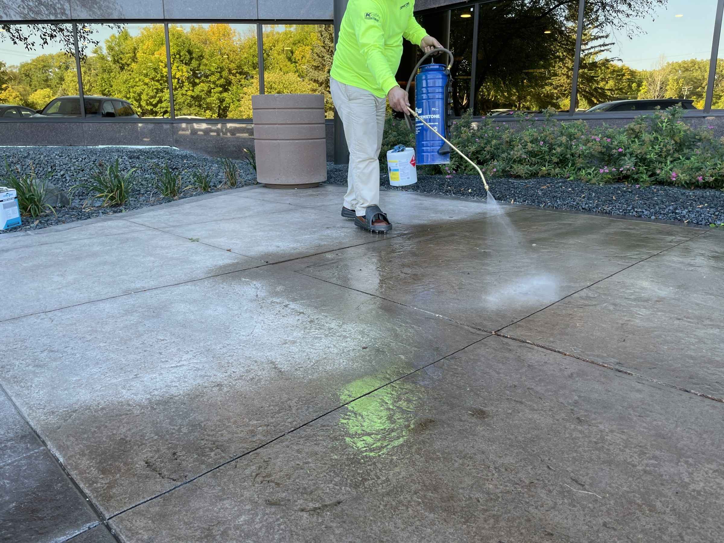 Worker with a pump sprayer applying sealer to a concrete patio outside a glass building